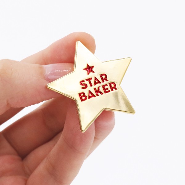 A hand holding a gold enamel plated Star Baker badge.