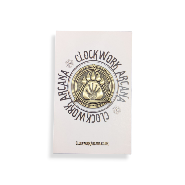 A gold antiqued custom 3D badge featuring an arcana symbol with a hand in a paw in a triangle within a circle. The pin badge is attached to a backing card with the Clockwork Arcana branding on it.
