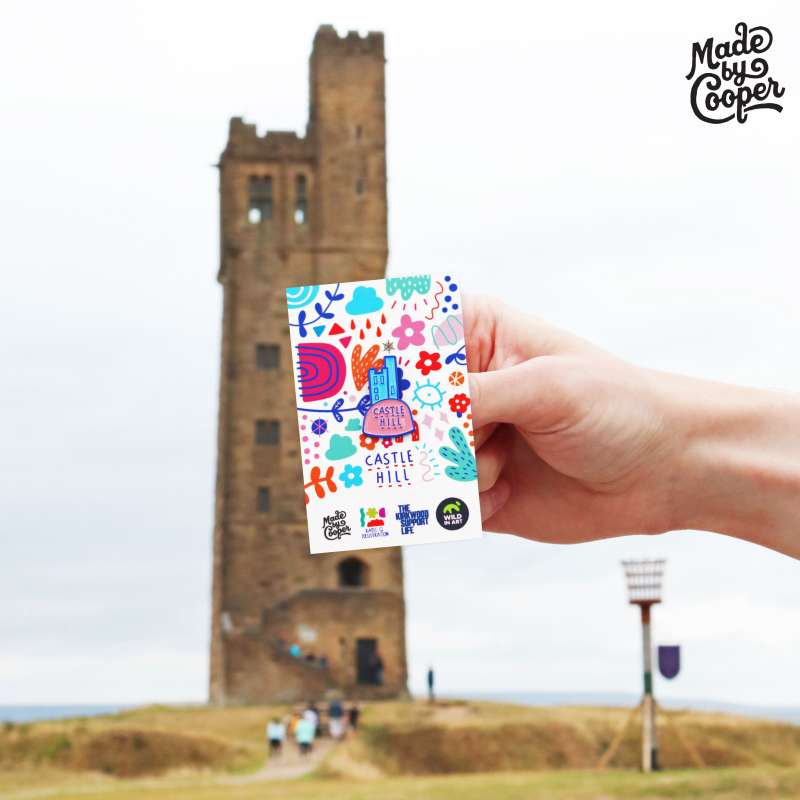 A stylised enamel badge of Castle Hill on a vibrant backing card being held up with Castle Hill in the backgorund.