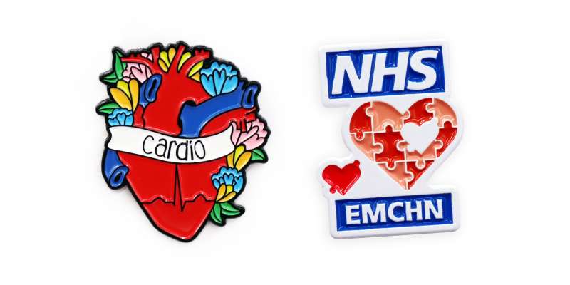 Two enamel pin badges. The first an anatomically correct heart surrounded by flowers with a banner that says 