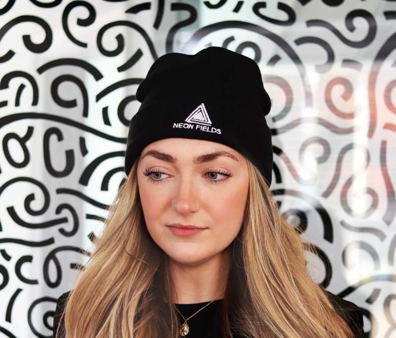 A women with long blonde hair wearing a black custom beanie with the Neon Fields logo embroidered on the front.