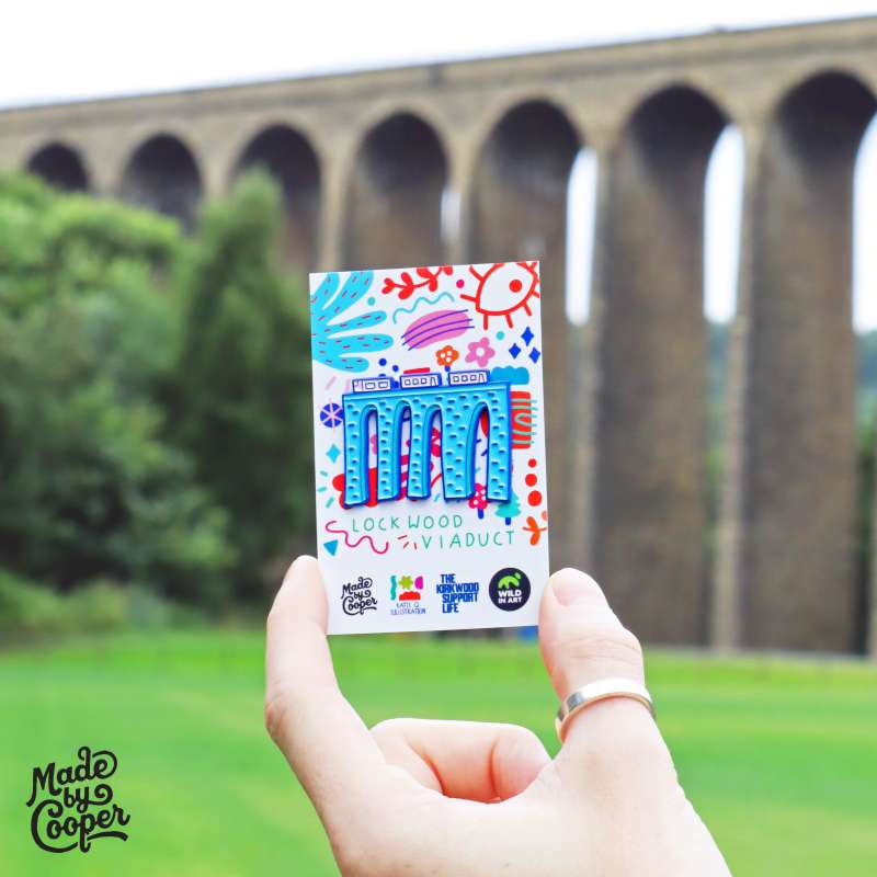 A stylised pin badge of Lockwood Viaduct on a colourful backing card being held up near Lockwood Viaduct.