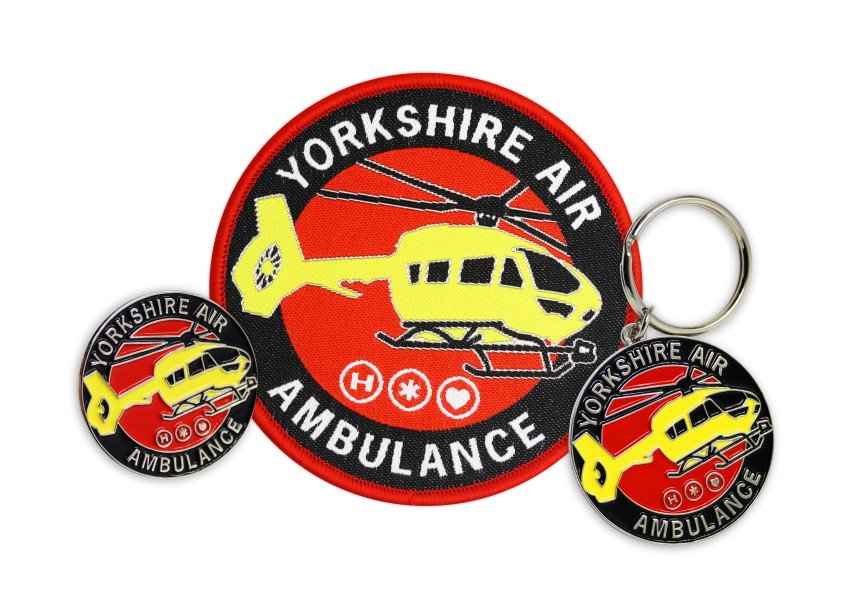A collection of Yorkshire Air Ambulance merch. A small soft enamel pin badge, a chunky soft enamel keyring, and a large woven patch, all sporting the new YAA red, black, and yellow logo and branding.