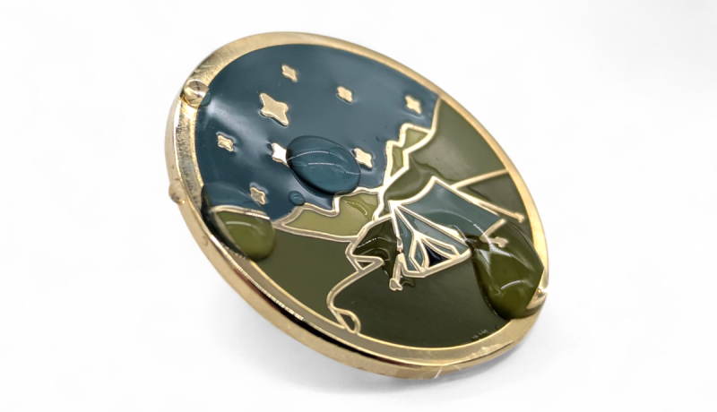 A profile view of a hard enamel pin badge with a campsite design. There are droplets of water on the front of the badge.