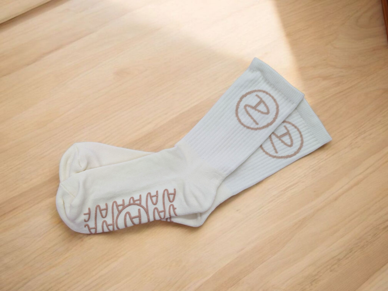 A pair of beige pilates socks laid on a wooden floor. The socks have a brown pattern at the top and a brown rubberised pattern on the sole for extra grip.