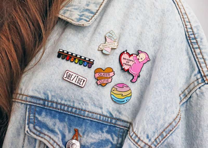 A light blue denim jacket with six LGBTQ+ related pin badges.