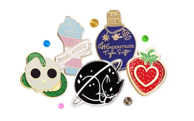 A collection of fun pin badges that people buy to express themselves. A skull and snake, trans rights hand, a sleeping cat, a Taylor Swift Wonderstruck bottle, and a cute strawberries.