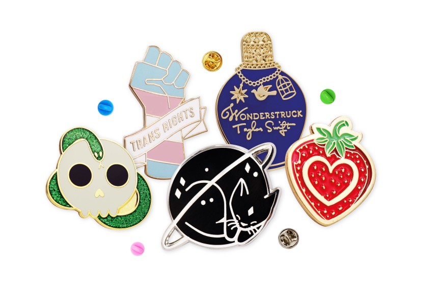 A collection of fun pin badges that people buy to express themselves. A skull and snake, trans rights hand, a sleeping cat, a Taylor Swift Wonderstruck bottle, and a cute strawberries.