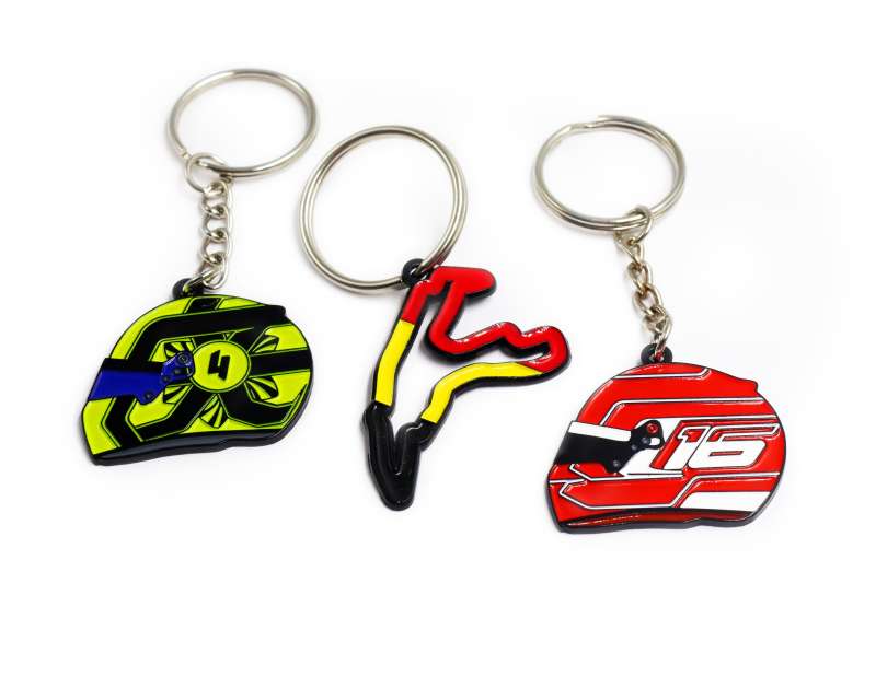 Three Formula 1 keyrings. A track map key from Spa Francochamps with the Belgium flag colours, and two helmet keyrings, one representing Lando Norris's #4 yellow helmet and another representing Charles Leclerc's #16 red helmet.