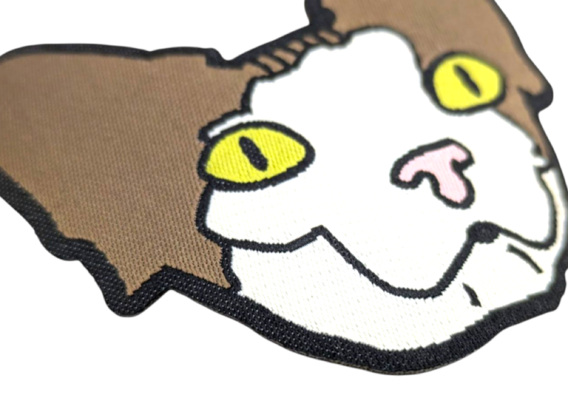 A close-up of a woven patch with heat-cut border in the shape of a car with white face, brown ears, and yellow eyes.