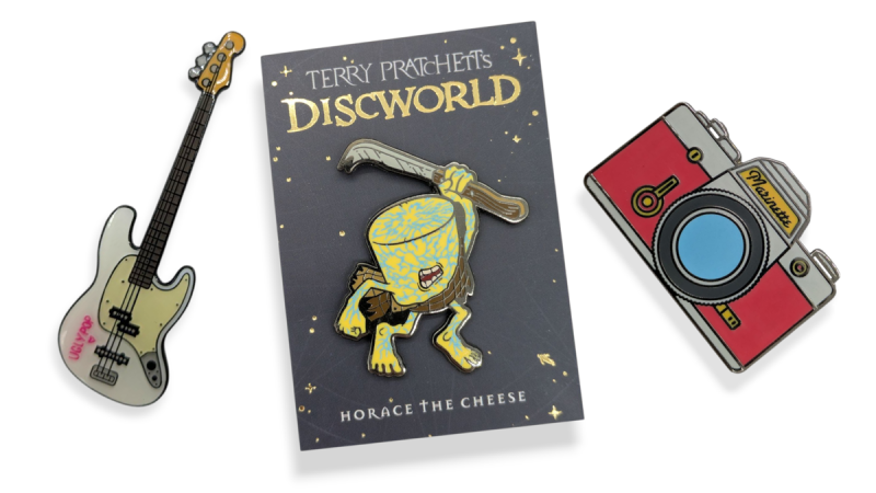 Three enamel pins with screen printing on them. An electric guitar, Horace the Cheese from Discworld, and a red camera.