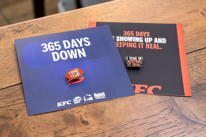 Two custom KFC anniversary pin badges displayed on branded backing cards on a wooden table, celebrating one year of service.