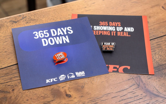 Two custom KFC anniversary pin badges displayed on branded backing cards on a wooden table, celebrating one year of service.
