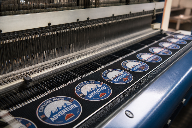 Industrial weaving machine producing custom woven patches in sequence, showing how detailed patch designs are manufactured.