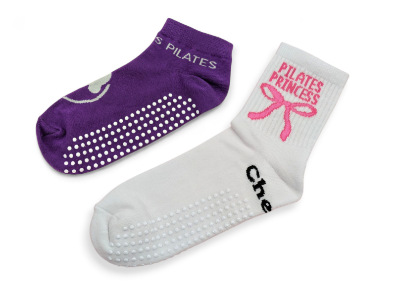 Two custom pilates socks. One a purple ankle height sock with white patterned grip, the other a white sports sock with white grip and a pink bow logo with the words