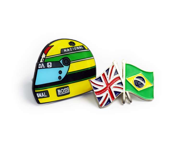 A helmet-shaped pin badge with Ayrton Senna's iconic helmet design, and a pin badge featuring the flags of the UK and Brazil representing the connection between Lewis Hamilton and Senna.