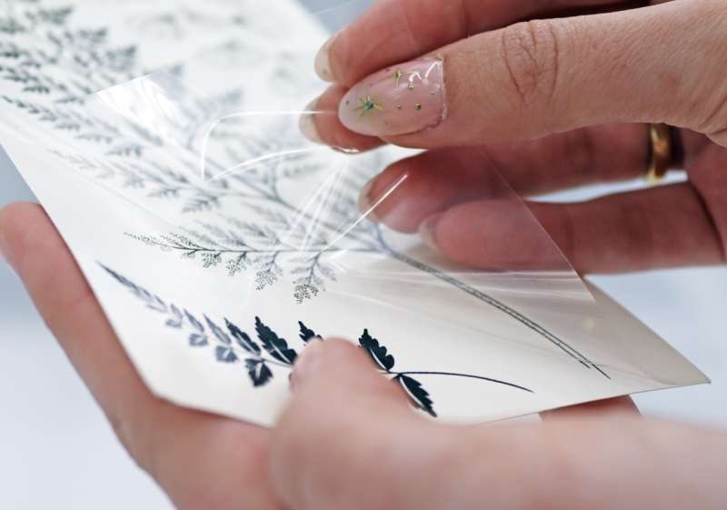 A female hand with fancy nails peels back the transparent film from a sheet of temporary tattoos.