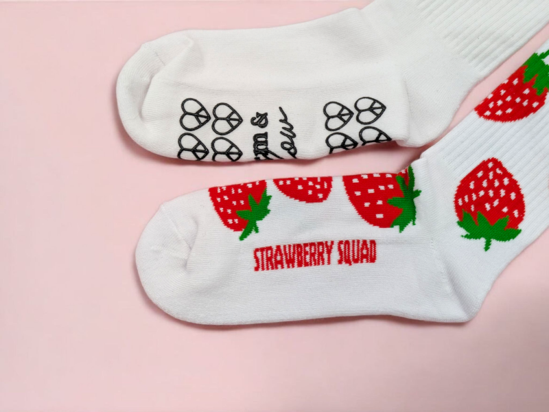 Two socks laid on a pink background. The top sock is a white pilates sock with a rubberised patterned sole, the bottom sock is a white sports socks with a strawberry pattern knitted in.