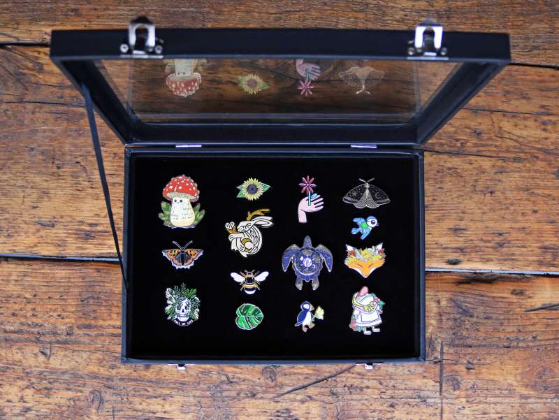 A black pin badge display case holding 16 nature related badges. The case is sat on a rustic wooden table.