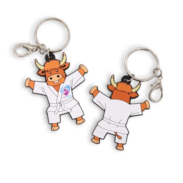 Custom PVC keyring featuring cartoon Highland cow character wearing a white judo gi with Judo Scotland logo, shown front and back with metal split ring and lobster clasp attachment.