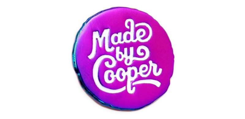 A white Made by Cooper logo on a rainbow plated round enamel pin badge.