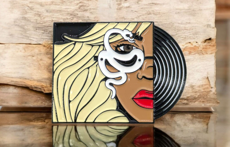 An illustrated pin badge of a record peeking out of it's sleeve. On the sleeve there is a blonde haired woman with bright red lip stick and a white snake in front of her eye.