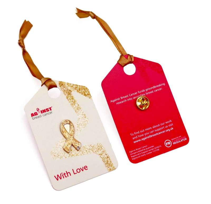 A golden breast cancer awareness ribbon pin badge on a beige backing card and brown ribbon. The back of the card is red.