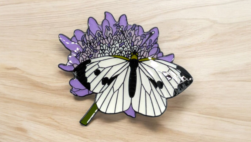 A beautifully designed pin badge of a butterfly sitting on a purple flower.
