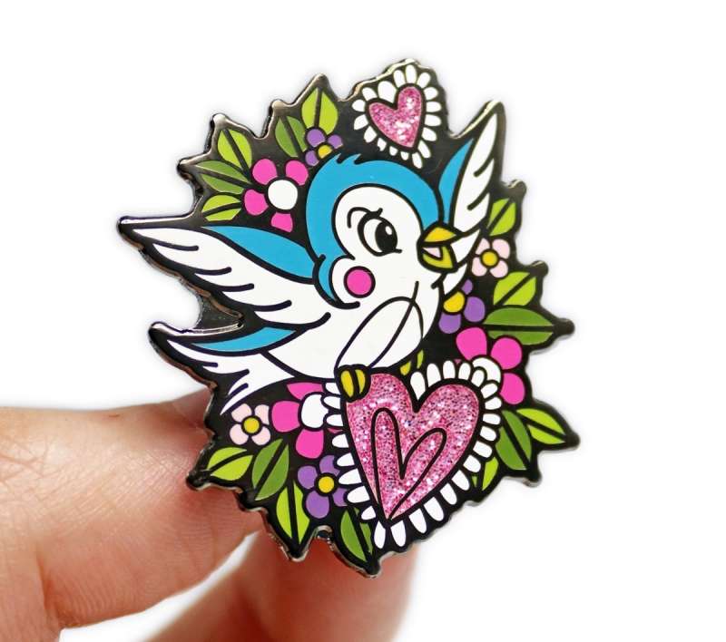 A stunning pin badge with a high-quality design of a cartoon swallow surrounded by love hearts and leaves.