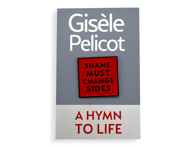 The book cover of A Hymn to Life by Gisele Pelicot featuring a bold red hard enamel “Shame Must Change Sides” pin badge mounted centrally on a grey and white cover design. The badge has raised black lettering and a glossy finish, creating strong contrast.
