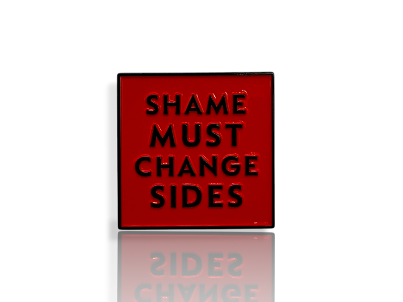 Close-up product photograph of the red hard enamel “Shame Must Change Sides” pin badge with raised black lettering. The square badge has a polished gloss surface and black metal edging, shown against a clean white background with a subtle reflection.