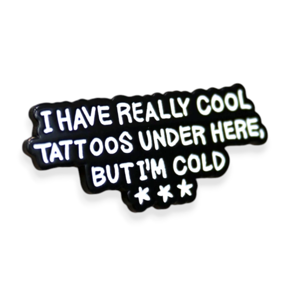 A custom enamel pin badge featuring the humorous phrase I have really cool tattoos under here but I’m cold, designed with bold white lettering on a black background and shaped text styling, ideal for artists, studios and playful custom pin merch ranges.