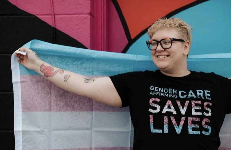 Stevie Brocksom rocking a black t-shirt with the slogan Gender Affirming Care Saves Lives, while holding a trans pride flag behind their back.