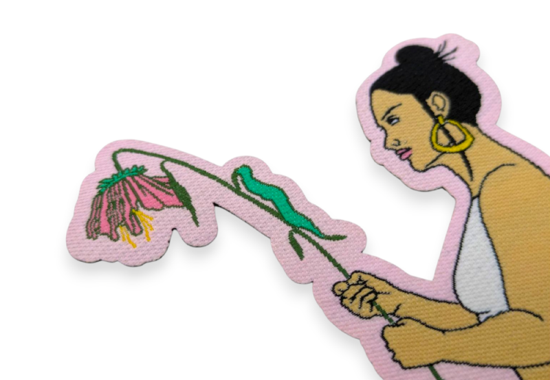 A close-up of a woven custom patch with a heat-cut border. The patch features a women with black hair holding a large but wilting flower.