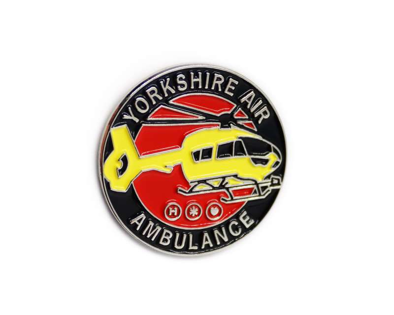 A soft enamel circular pin badge featuring the yellow helicopter of the Yorkshire air ambulance on a red and black background. The words Yorkshire Air Ambulance encircle the badge.